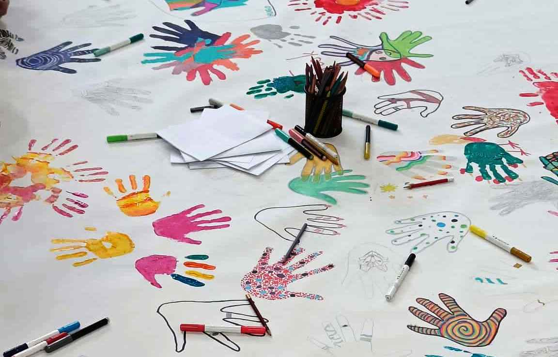 dziecięce dłonie poodbijane kolorowymi farbami na białym papierze, rozrzucone flamastry i kredki