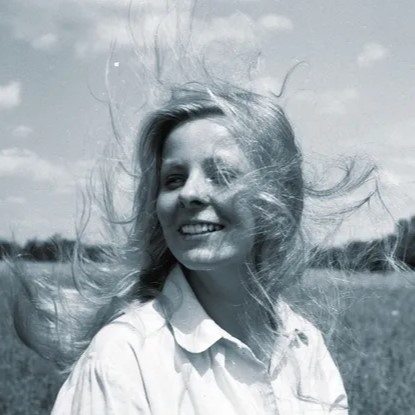 Czarno-biała fotografia Anny Nehrebeckiej. Aktorka w jasnej koszuli stoi na polu w słoneczny dzień, z rozwianymi na wietrze włosami, w tle widoczne niebo z chmurami.