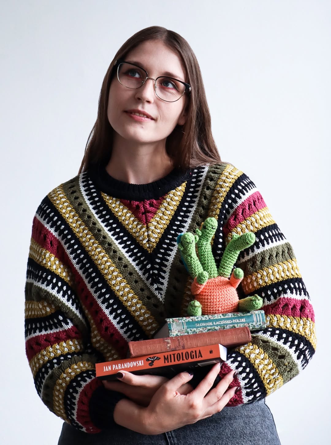 Zdjęcie Roksany Łajkosz, ma na sobie kolorowy sweter, rozpuszczone włosy, okulary. W rękach trzyma książki i mitologiczną zabawkową hydrę zrobioną na szydełku. 