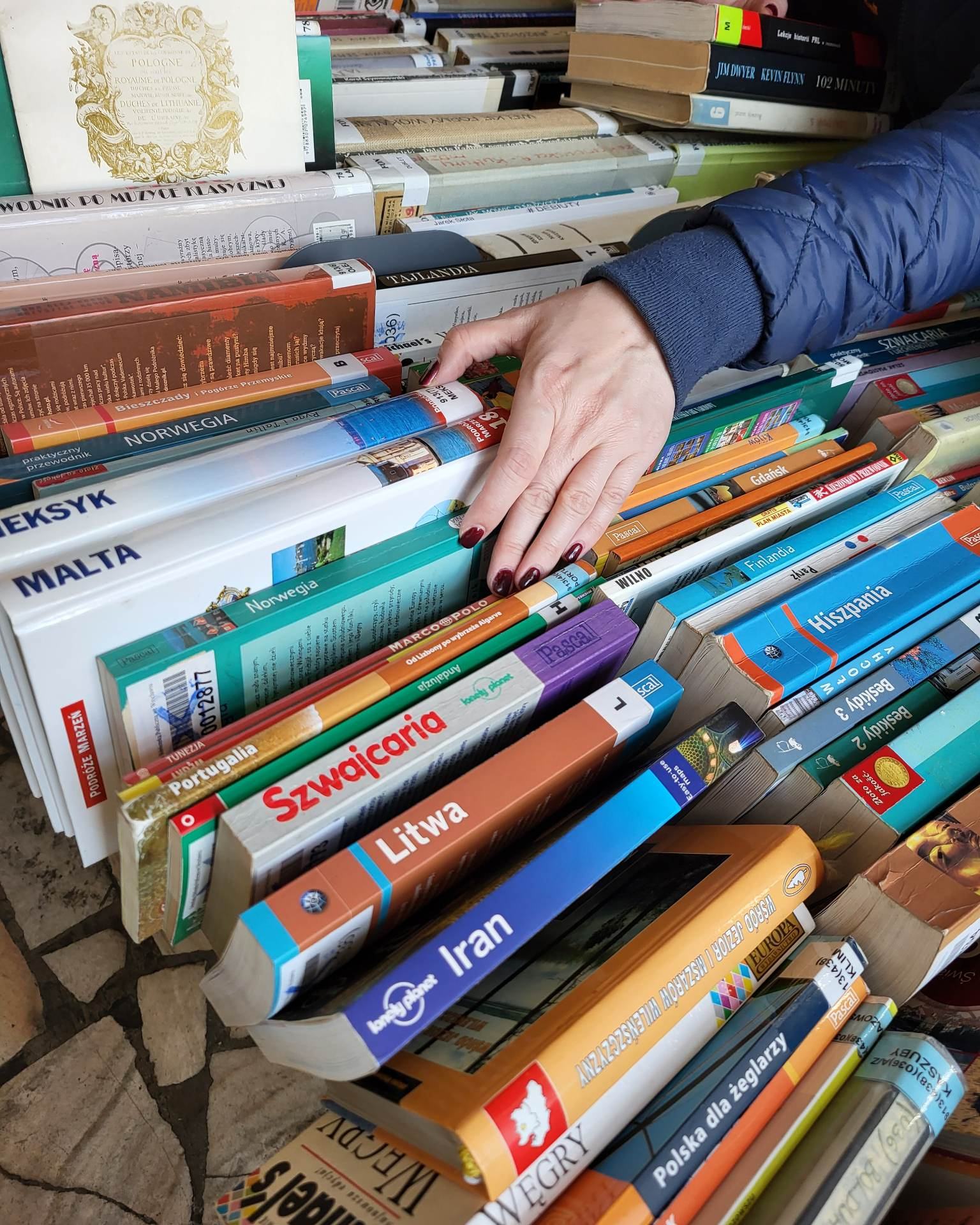 Zbliżenie na rękę osoby przesuwającej przewodniki i książki podróżnicze ułożone w rzędach. Wśród widocznych tytułów znajdują się publikacje o krajach takich jak Malta, Norwegia, Szwajcaria, Litwa i Iran.