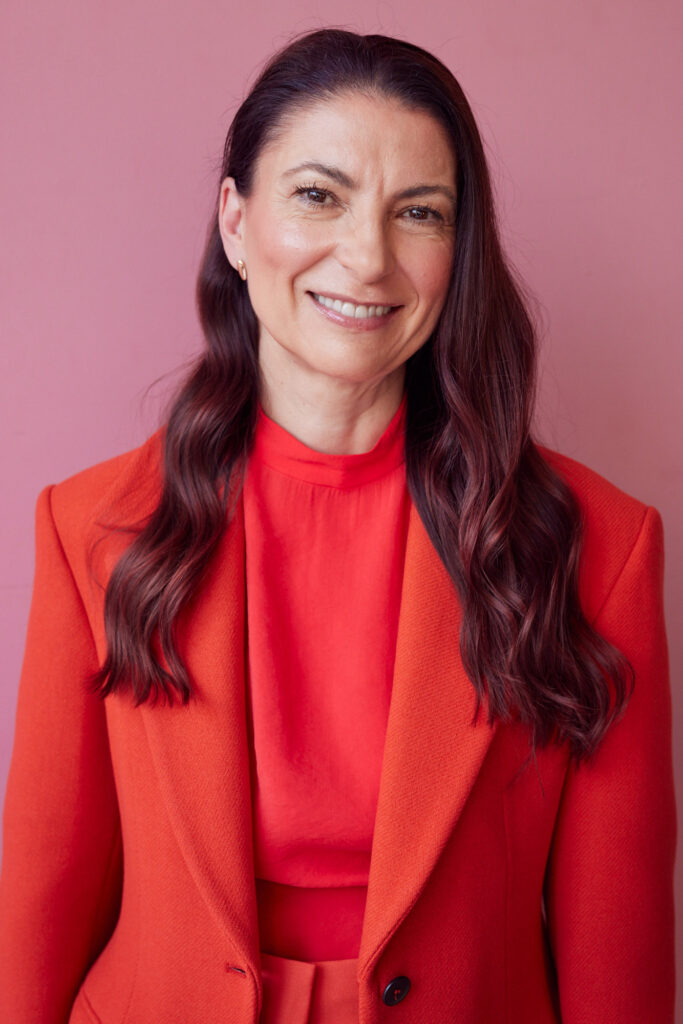 Na zdjęciu widoczna jest ciemnowłosa kobieta w czerwonej marynarce na różowym tle.