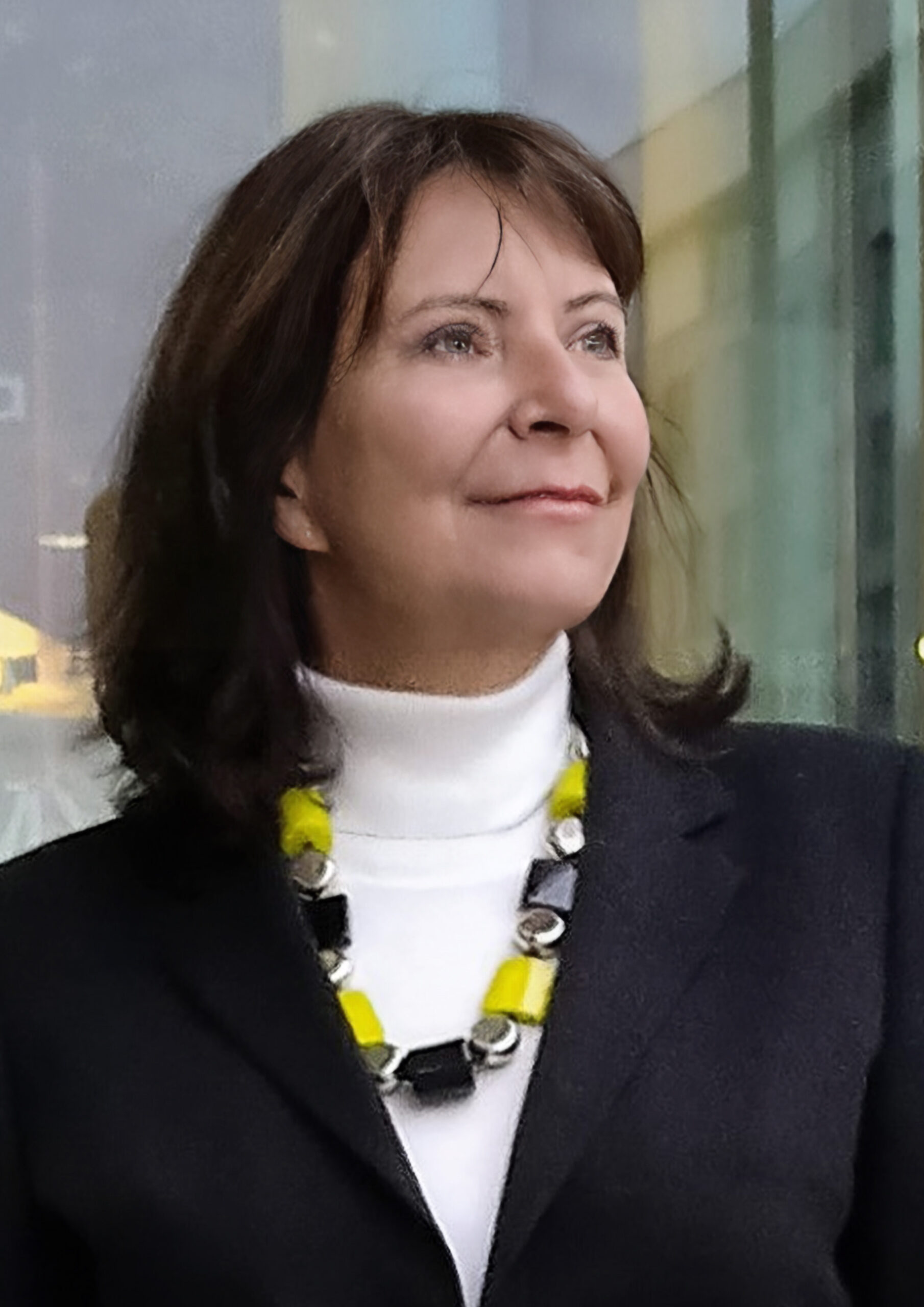 dr Izabella Starzec, portret kobiety w czarnej marynarce, dużych koralach i rozpuszczonych włosach, w tle lekko rozmyty obraz miasta w szybach