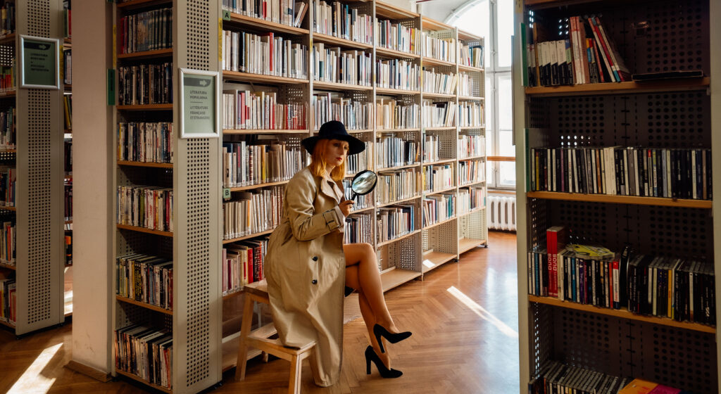 pani detektyw w długim płaszczu siedzi w Bibliotece Romańskiej między regałami książek
