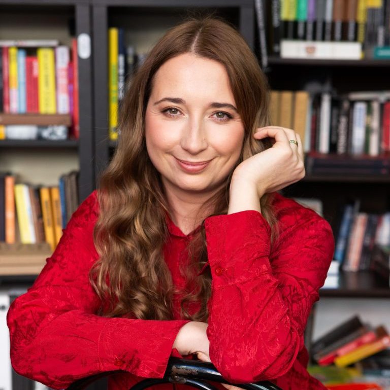 Marta Matuszczak w czerwonej sukience siedzi na krześle, za nią regały z książkami.