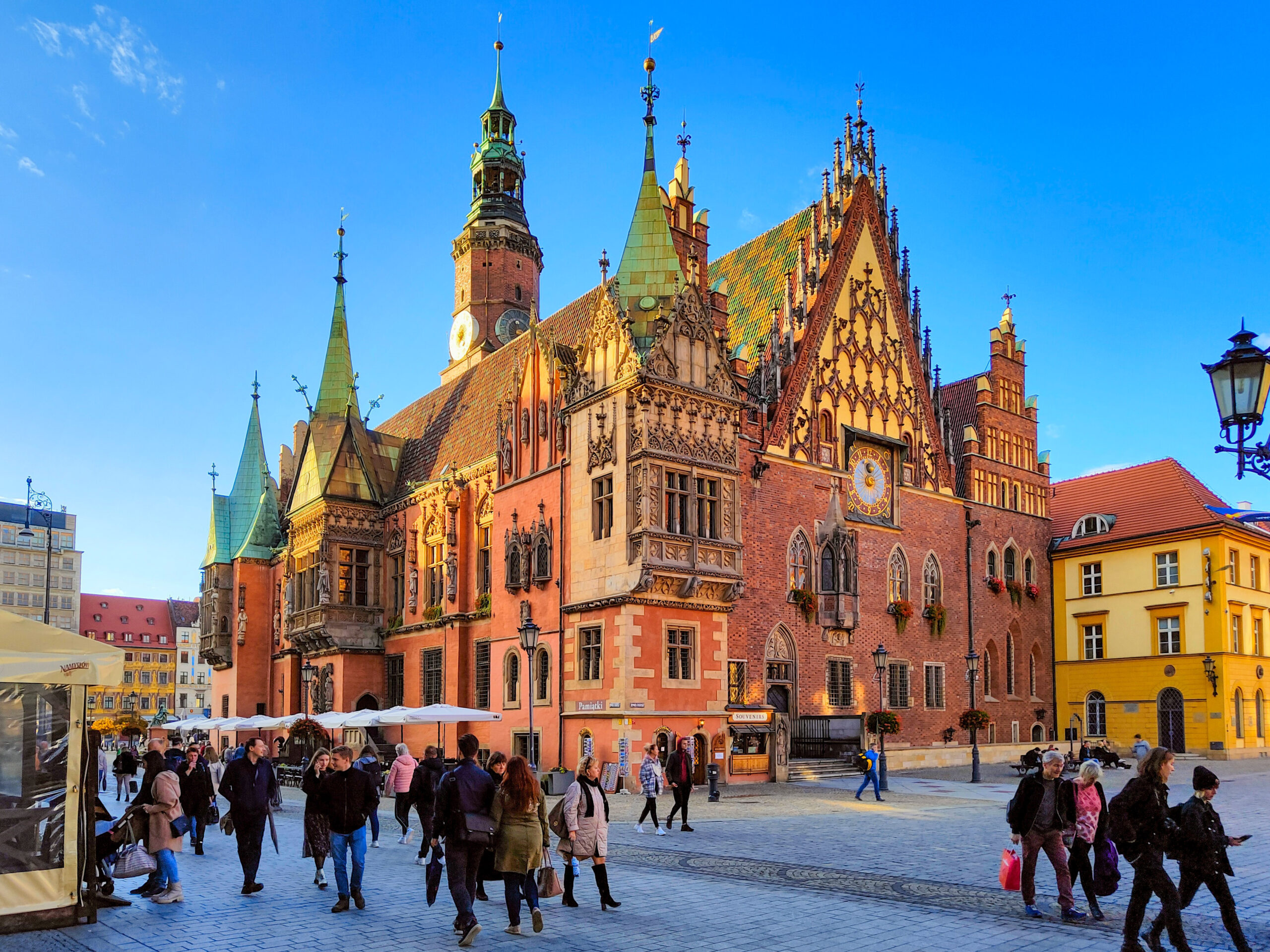 Historyczny ratusz na wrocławskim Rynku w słoneczny dzień, z licznymi przechodniami spacerującymi po placu.