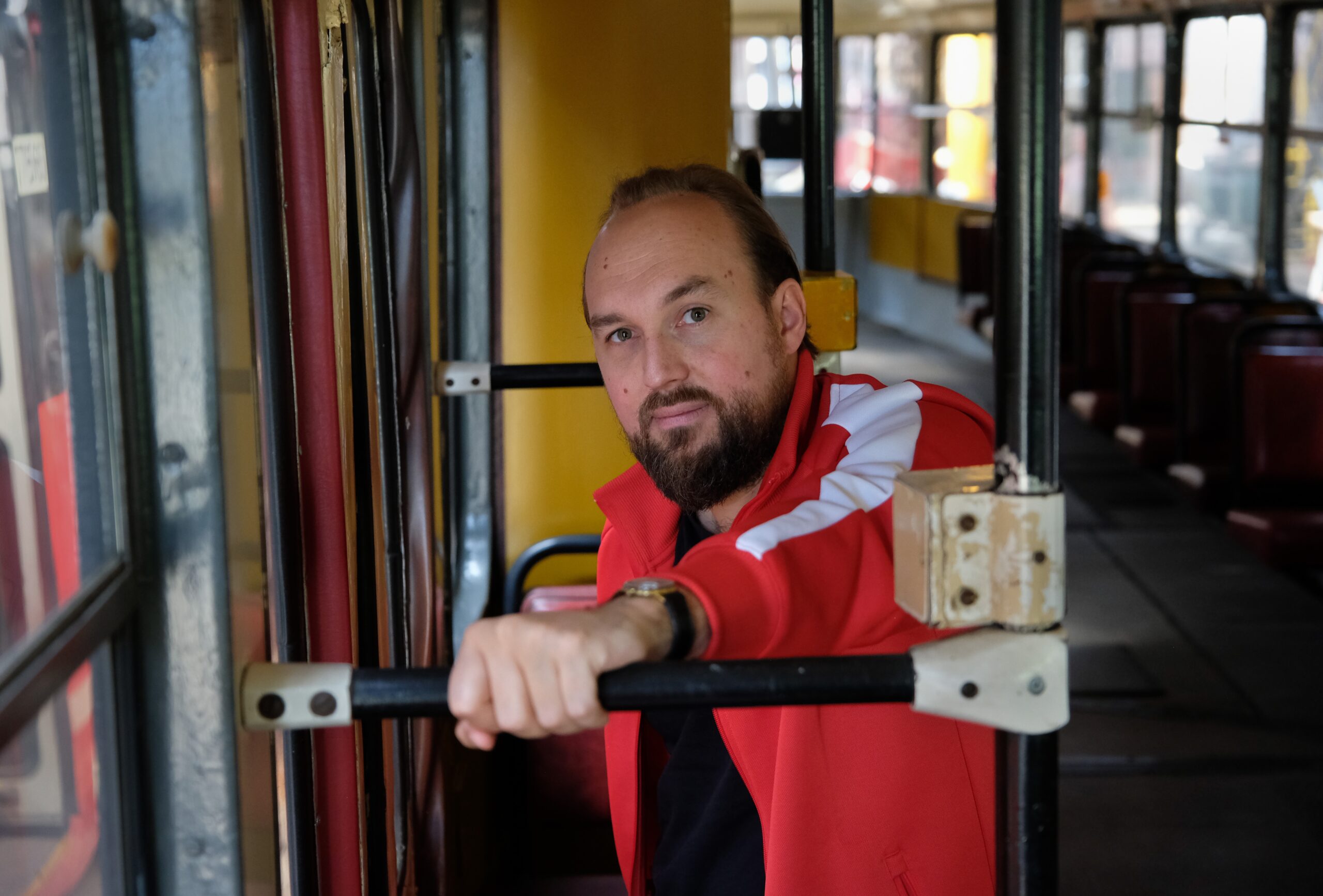 Na fotografii widoczny jest mężczyzna w czerwonej bluzie, z brodą, który znajduje się w tramwaju.
