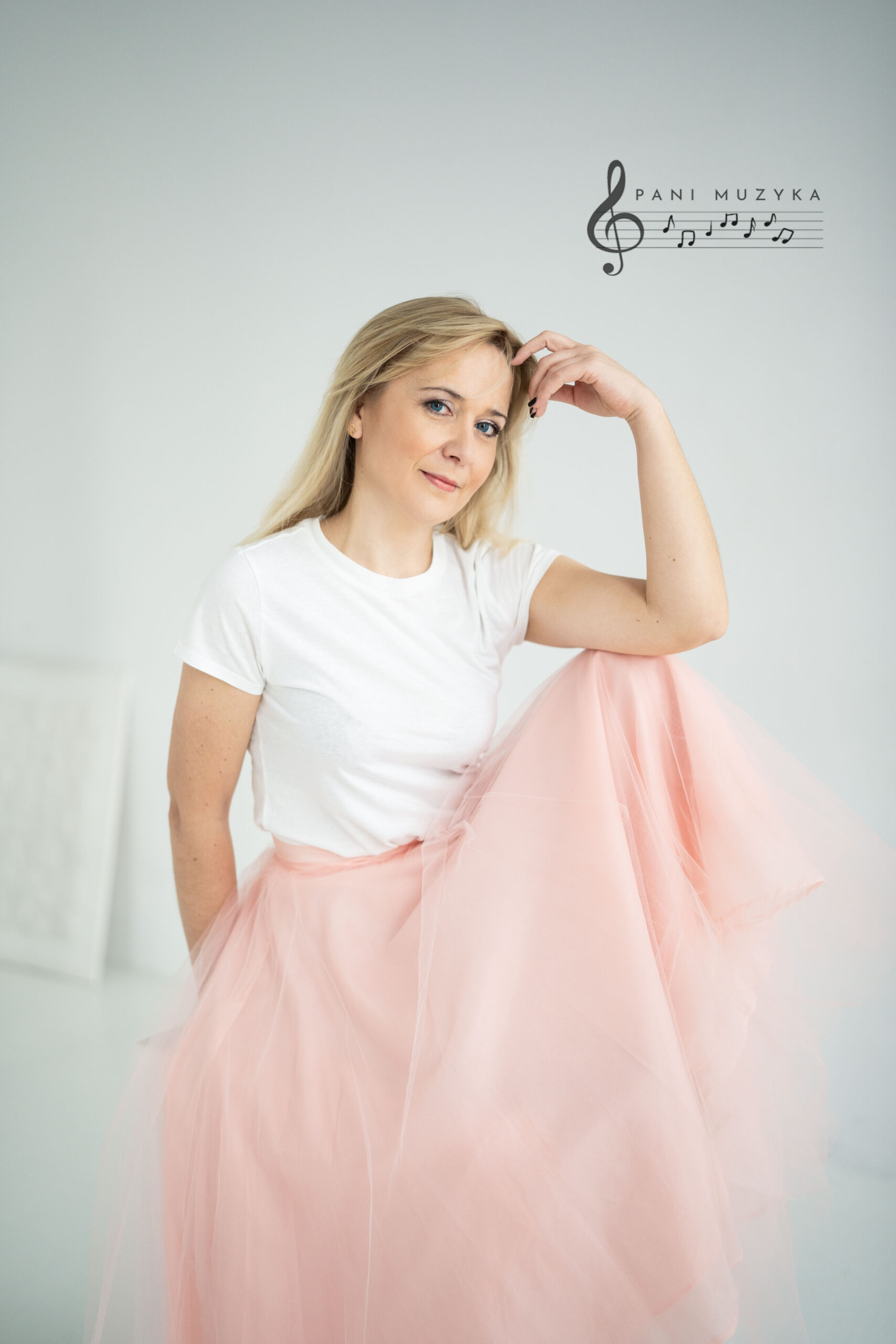 Portret kobiety ubranej w biały t-shirt i obszerną, jasnoróżową tiulową spódnicę. Kobieta siedzi, patrząc łagodnie w stronę obiektywu, z jedną ręką uniesioną przy czole. W prawym górnym rogu znajduje się czarny logotyp przedstawiający klucz wiolinowy oraz pięciolinię z napisem „PANI MUZYKA”.