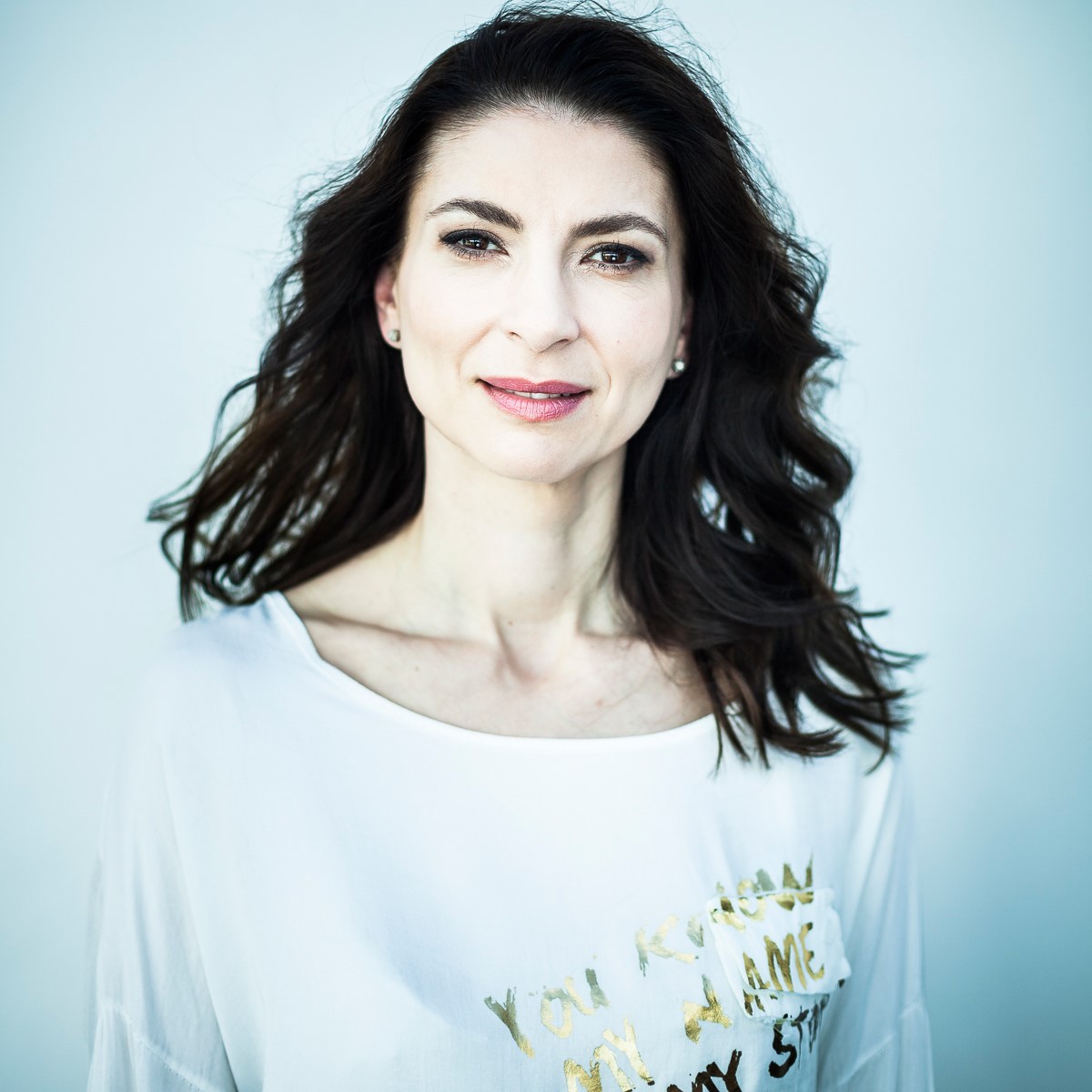 Na fotografii widoczna jest czarnowłosa kobieta ubrana w białą koszulę na jasnoniebieskim tle.