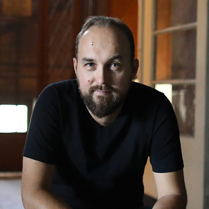 Zbigniew Rokita w czarnym podkoszulku, z zarostem, w tle przestrzeń klatki schodowej