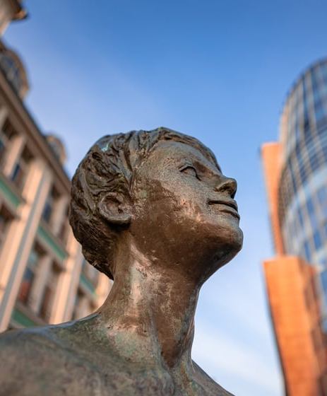 Zbliżenie na brązową rzeźbę przedstawiającą nagą sylwetkę o uniesionej głowie, ustawioną na tle miejskiej zabudowy i nowoczesnego przeszklonego budynku w słoneczny dzień.