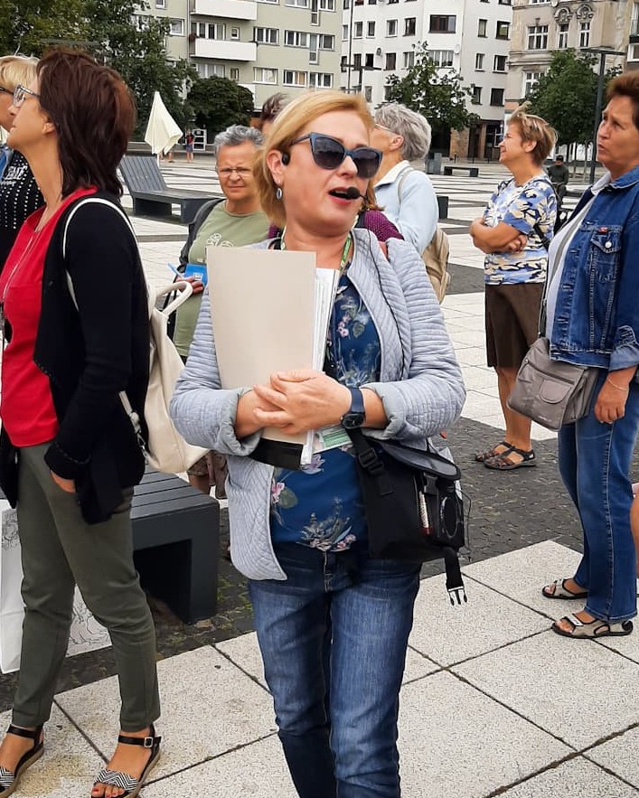 Krystyna Lasota-Kotowicz, kobieta z blond włosami, w ciemnych okularach, z mikrofonem i notatkami w rękach prowadzi wycieczkę przez Nowy Targ we Wrocławiu