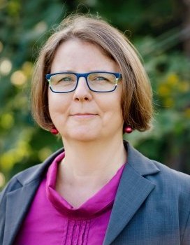 kobieta ma włosy za ucho, okulary, w tle zieleń, fioletowa bluzka i niebieska marynarka