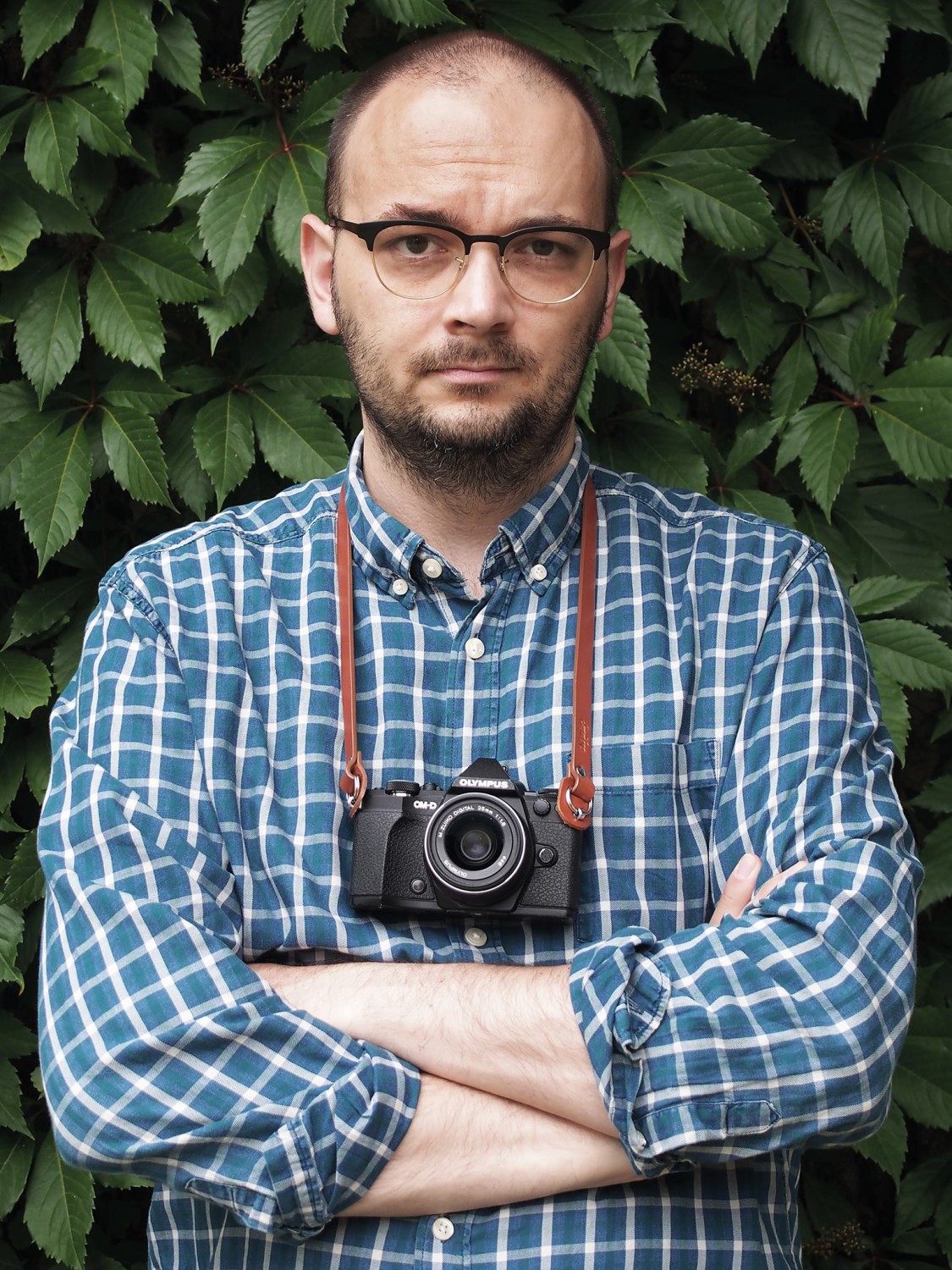 Na zdjęciu widoczny jest mężczyzna z aparatem fotograficznym i w niebieskiej koszuli w kratę.