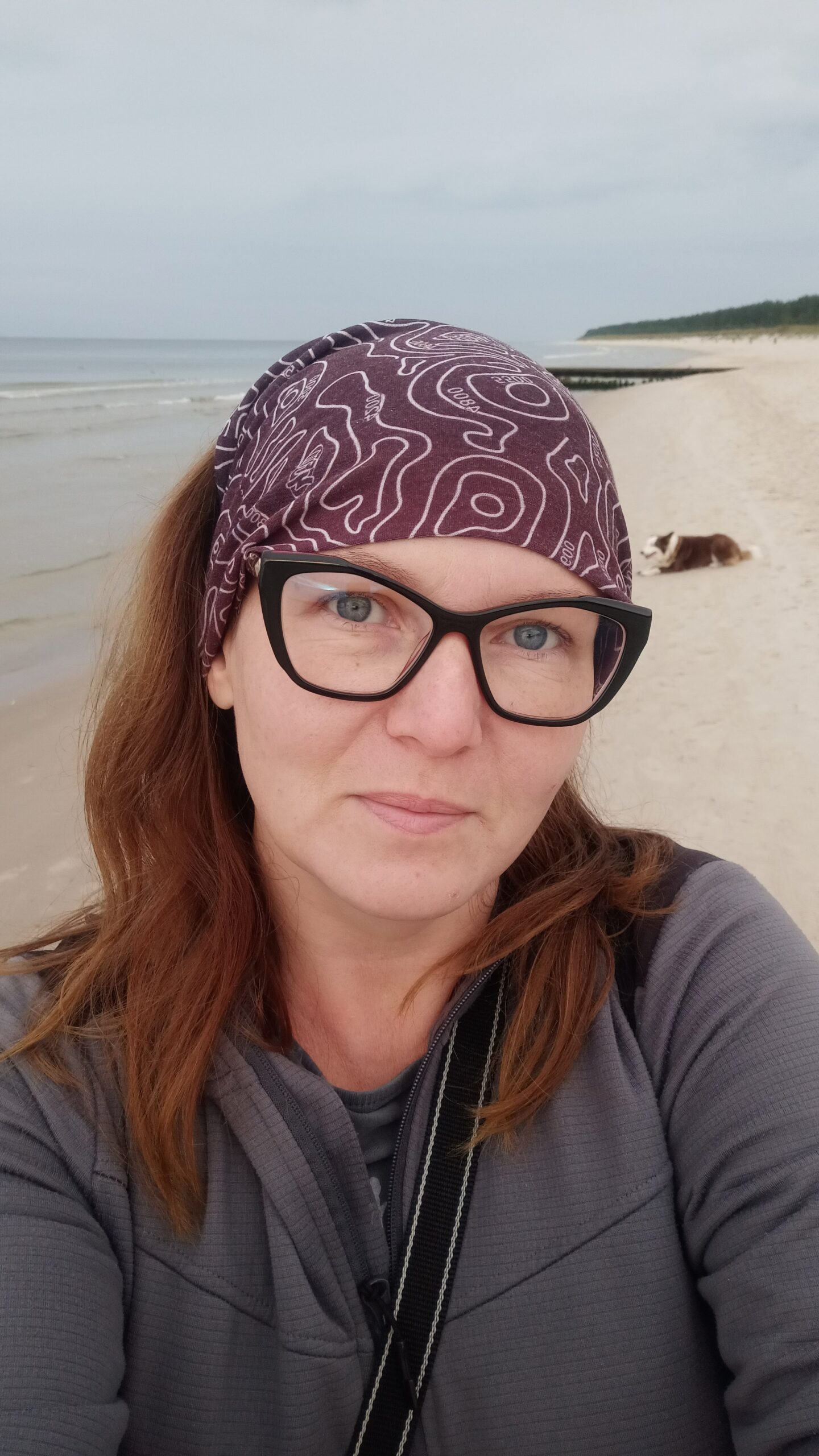 Kobieta w okularach, szarej bluzie i bordowej chuście na tle plaży 