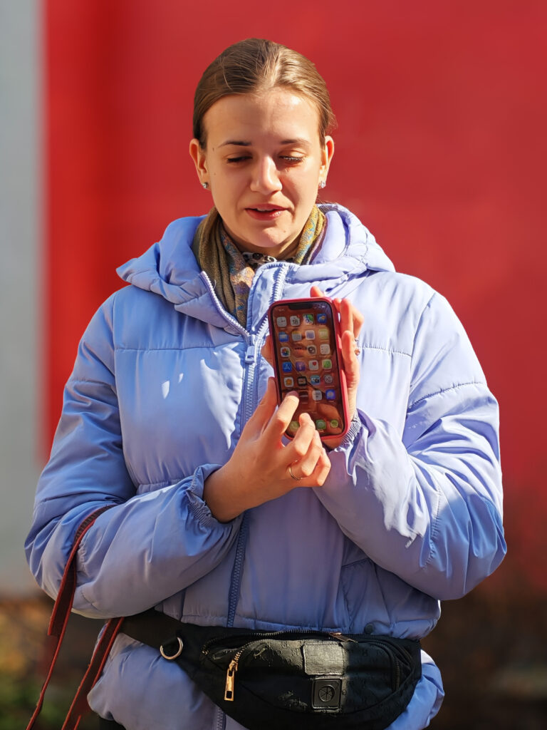 Niewidoma kobieta wskazuje palcem na aplikacje w telefonie. Z tyłu czerwona ściana. 