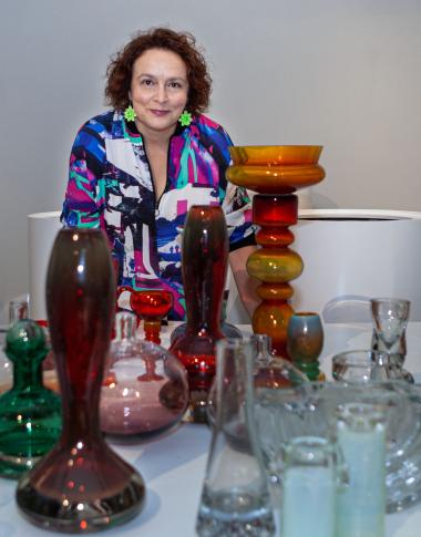 dr Barbara Banaś stoi za stołem zastawionym szkłem artystycznym, zdjęcie jest kolorowe i podkręca artystyczny charakter zarówno przedmiotów, jak i kobiety