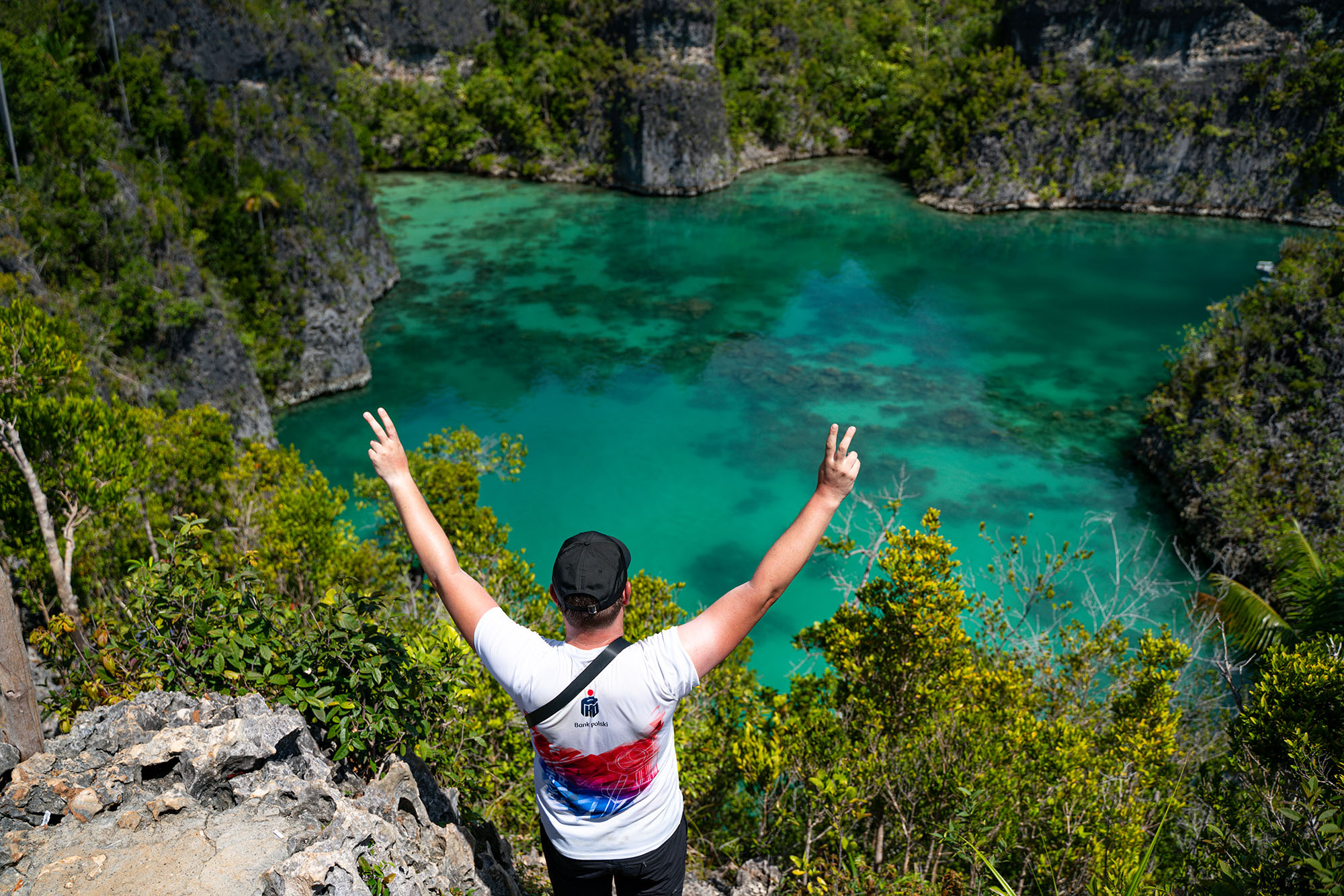 mężczyzna w białej koszulce i czarnej czapce z daszkiem z uniesionymi rękami odwrócony tyłem  na tle egzotycznego krajobrazu