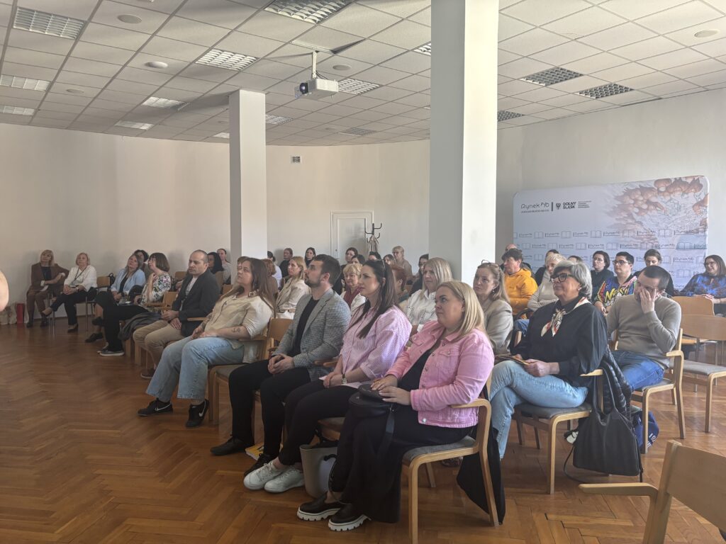 Publiczność siedząca na krzesłach w jasnej sali konferencyjnej, zwrócona w stronę niewidocznej prelegentki lub prelegenta; w tle filary, projektor pod sufitem i plansza wydarzenia