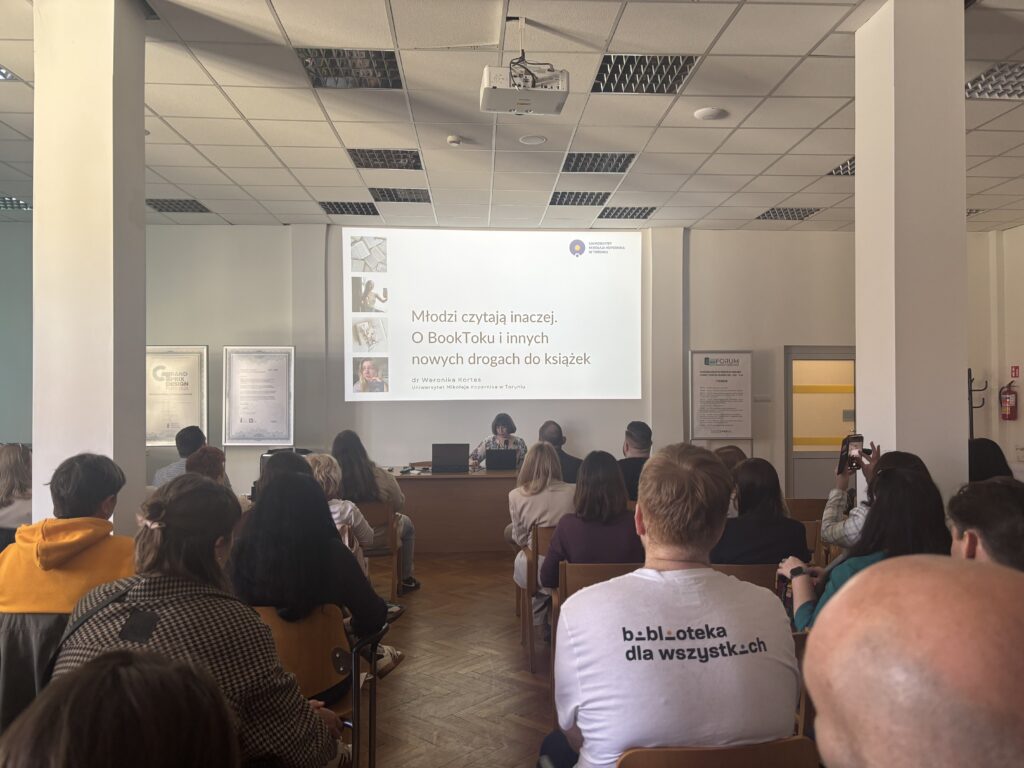 Widok od tyłu na uczestników konferencji słuchających wystąpienia; na ścianie wyświetlona prezentacja pt. „Młodzi czytają inaczej. O BookToku i innych nowych drogach do książek