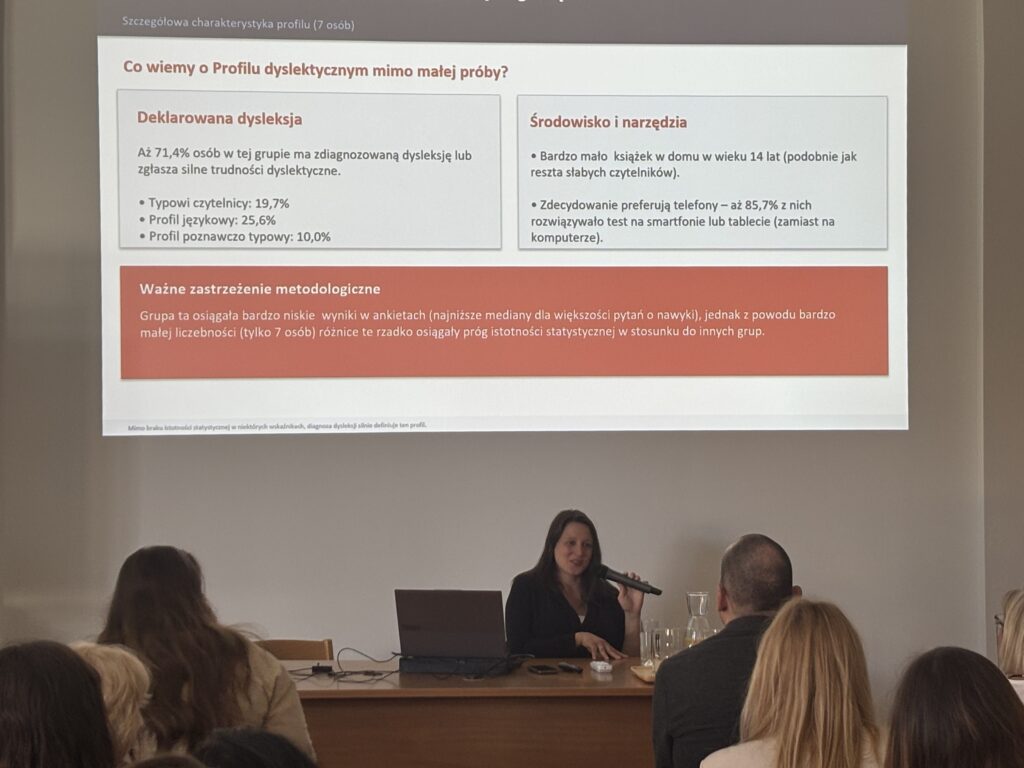 prelegentka siedząca przy stole z laptopem i mikrofonem, przed publicznością; za nią slajd z wynikami badania dotyczącego profilu dyslektycznego