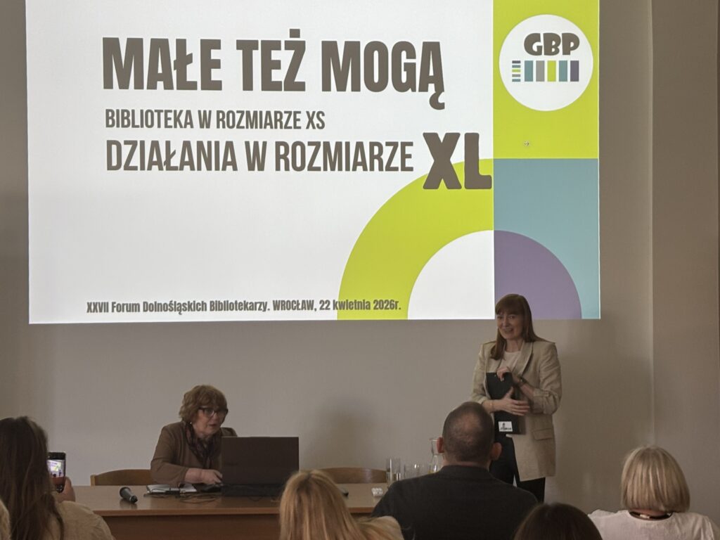Dwie osoby prowadzące prezentację przy stole; na ekranie slajd „Małe też mogą. Biblioteka w rozmiarze XS, działania w rozmiarze XL” z logo GBP i informacją o forum bibliotekarzy we Wrocławiu