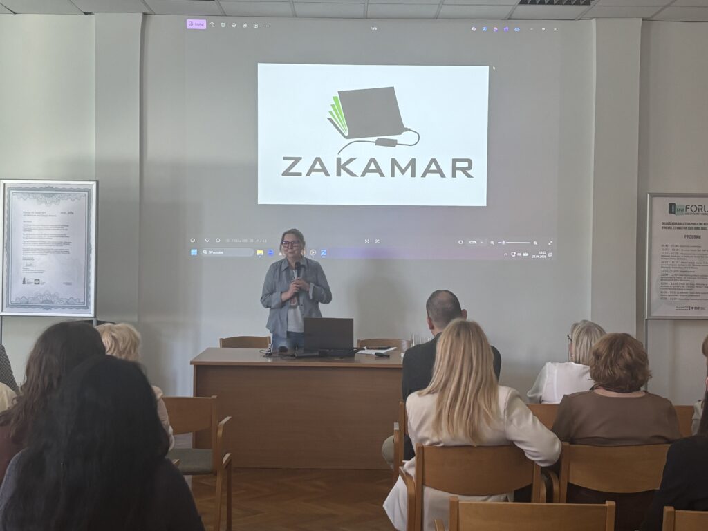 Prelegentka stojącA przy stole z laptopem i mikrofonem, przed publicznością; na ekranie slajd z logo „ZAKAMAR"
