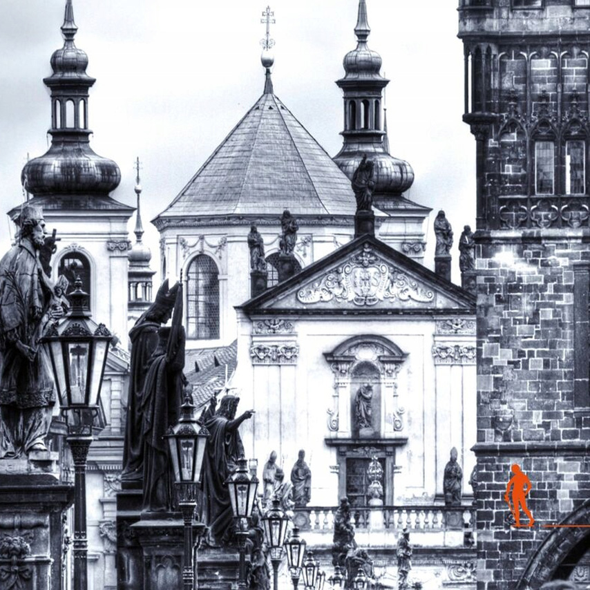 Czarno-biała fotografia przedstawiająca zabytkowy most wyłożony kamieniem, po którego obu stronach stoją rzeźby na wysokich cokołach oraz stylowe latarnie. W tle widoczne są ozdobne budynki sakralne z kopułami, wieżami i bogatymi detalami architektonicznymi. Po prawej stronie znajduje się fragment masywnej, kamiennej wieży.