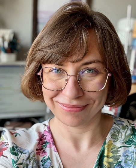 Osoba z krótkimi, jasnobrązowymi włosami, ubrana w bluzkę w kwiatowy wzór, sfotografowana w pomieszczeniu z widocznym w tle biurkiem i akcesoriami