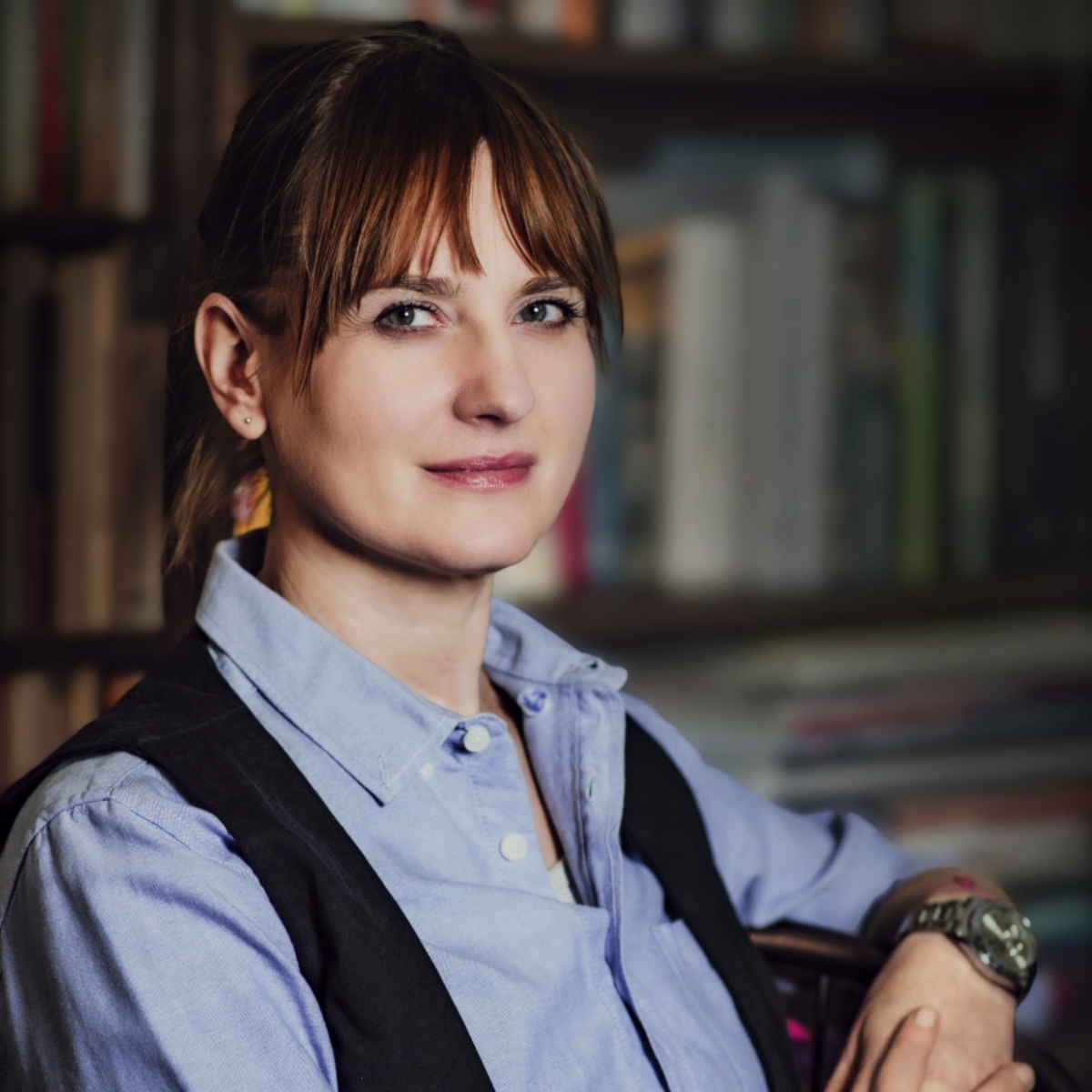 Portret kobiety w niebieskiej koszuli, ma splecione dłonie, w tle znajduje się regał z książkami