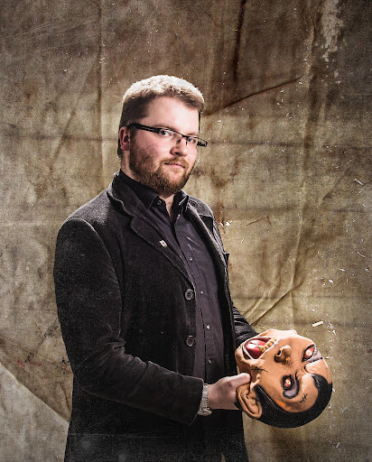 Osoba w ciemnym stroju stoi na tle postarzonej, teksturowanej powierzchni, trzymając w dłoniach dużą teatralną maskę o wyrazistej, dramatycznej mimice.