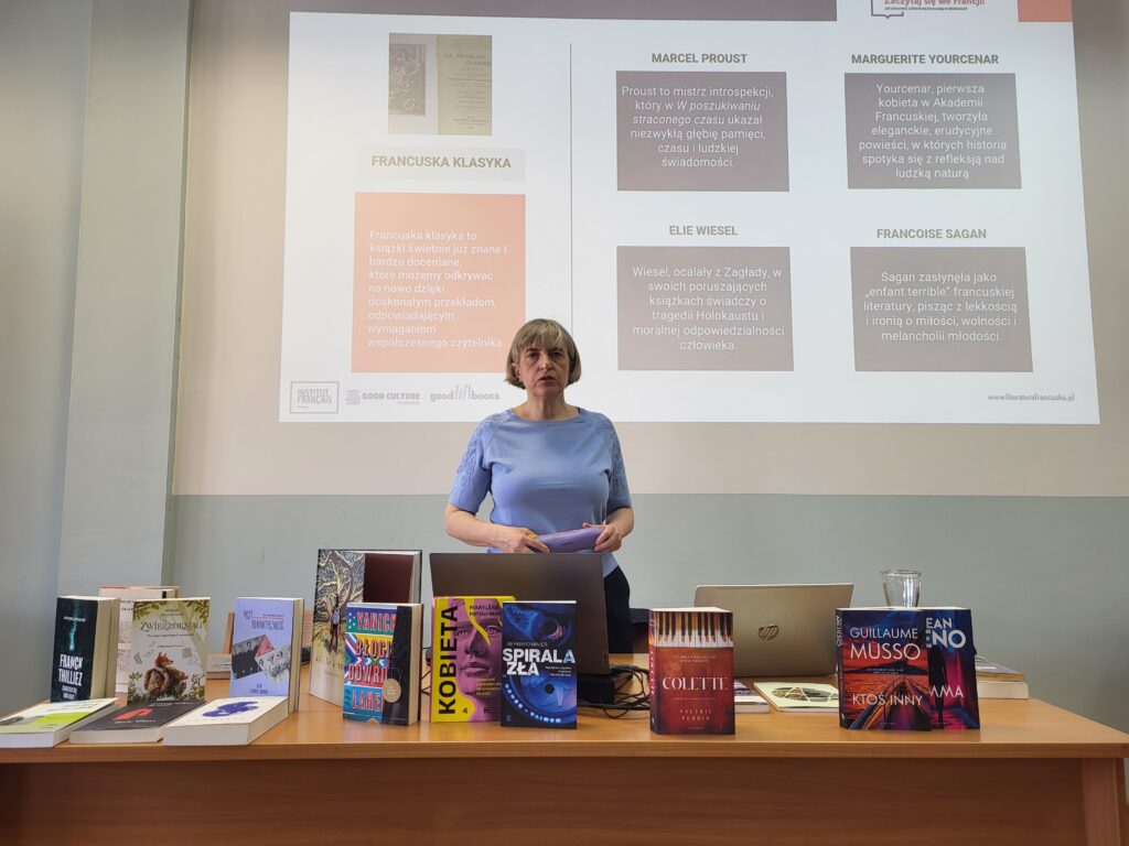 Osoba stojąca za stołem prezentacyjnym mówi do publiczności. Przed nią ustawione są książki i laptop. W tle wyświetlany jest slajd zatytułowany „Francuska klasyka” z nazwiskami autorów, m.in. Marcel Proust, Marguerite Yourcenar, Elie Wiesel i Françoise Sagan.