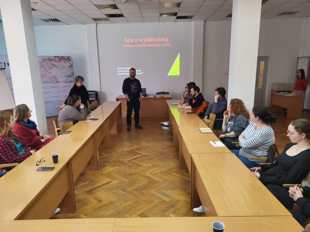 Spotkanie warsztatowe w sali wykładowej: uczestnicy siedzą przy ustawionych w podkowę stołach, a osoba stojąca pośrodku prowadzi zajęcia. W tle widoczny jest slajd z napisem „Gra z wyobraźnią, czyli co trzeba wiedzieć o RPG”.