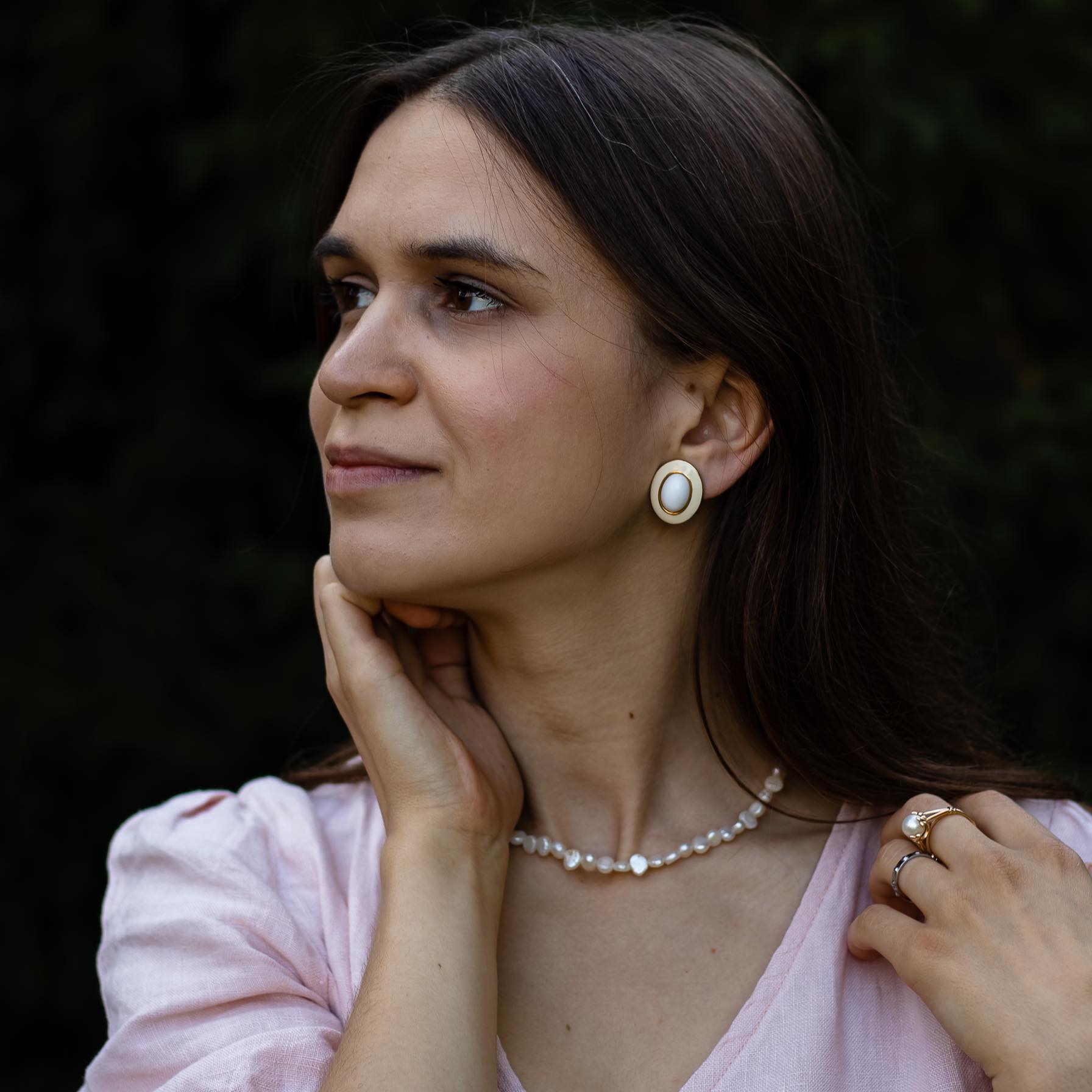 portret kobiety, profil, trzyma dłoń na twarzy, ma perłową biżuterię, ciemne włosy i różową sukienkę