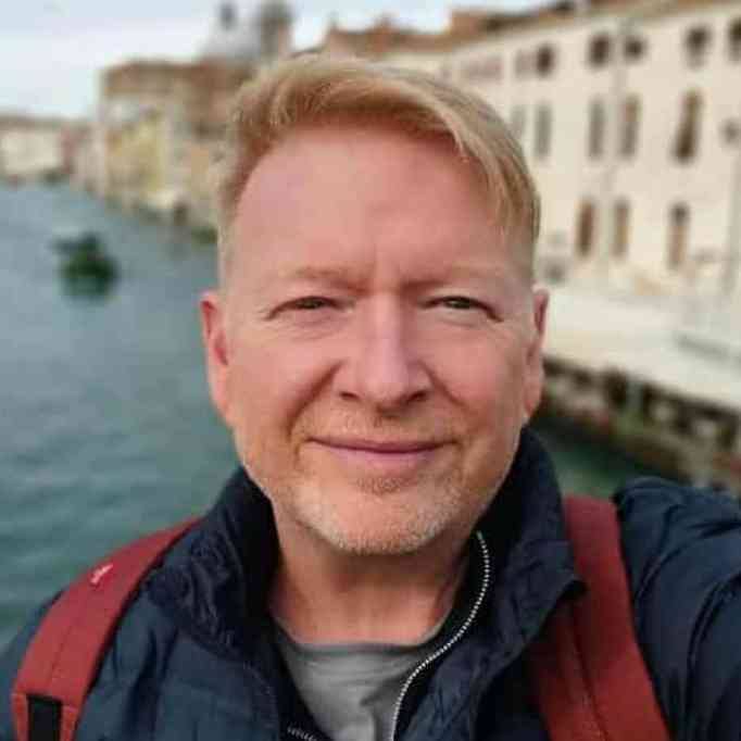 Richard Darcy, portret mężczyzny w średnim wieku, w tle rzeka i lekko rozmyta architektura. Mężczyzna uśmiecha się lekko i patrzy w obiektyw. 