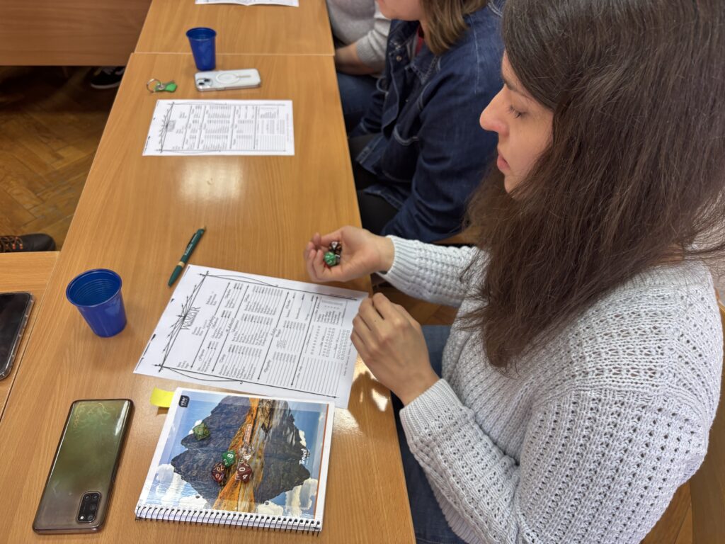 Uczestniczka siedząca przy stole trzyma zieloną kostkę do gry. Przed nią leży karta z tabelą, długopis, telefon oraz notes z ilustracją.