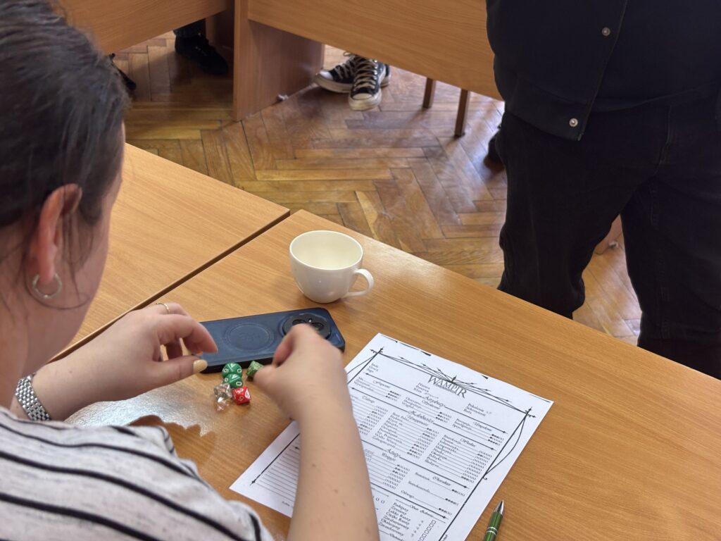 Uczestnik siedzący przy stole rzuca lub układa kolorowe kostki do gry obok karty z tabelą. Na stole znajduje się również filiżanka i telefon.