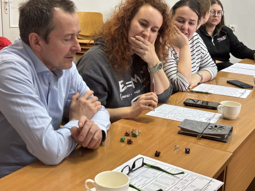 Grupa uczestników siedzących przy stole, pochylonych nad kartami z tabelami. Na stole leżą kostki do gry, długopisy, telefon, etui na okulary oraz filiżanka.