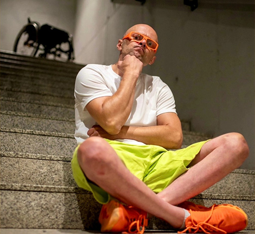 Mężczyzna siedzący na schodach w swobodnej pozie, opierający brodę na dłoni. Ma na sobie biały t-shirt, neonowo zielone szorty oraz pomarańczowe buty i okulary. W tle widać wnętrze budynku z betonowymi schodami i poręczą.