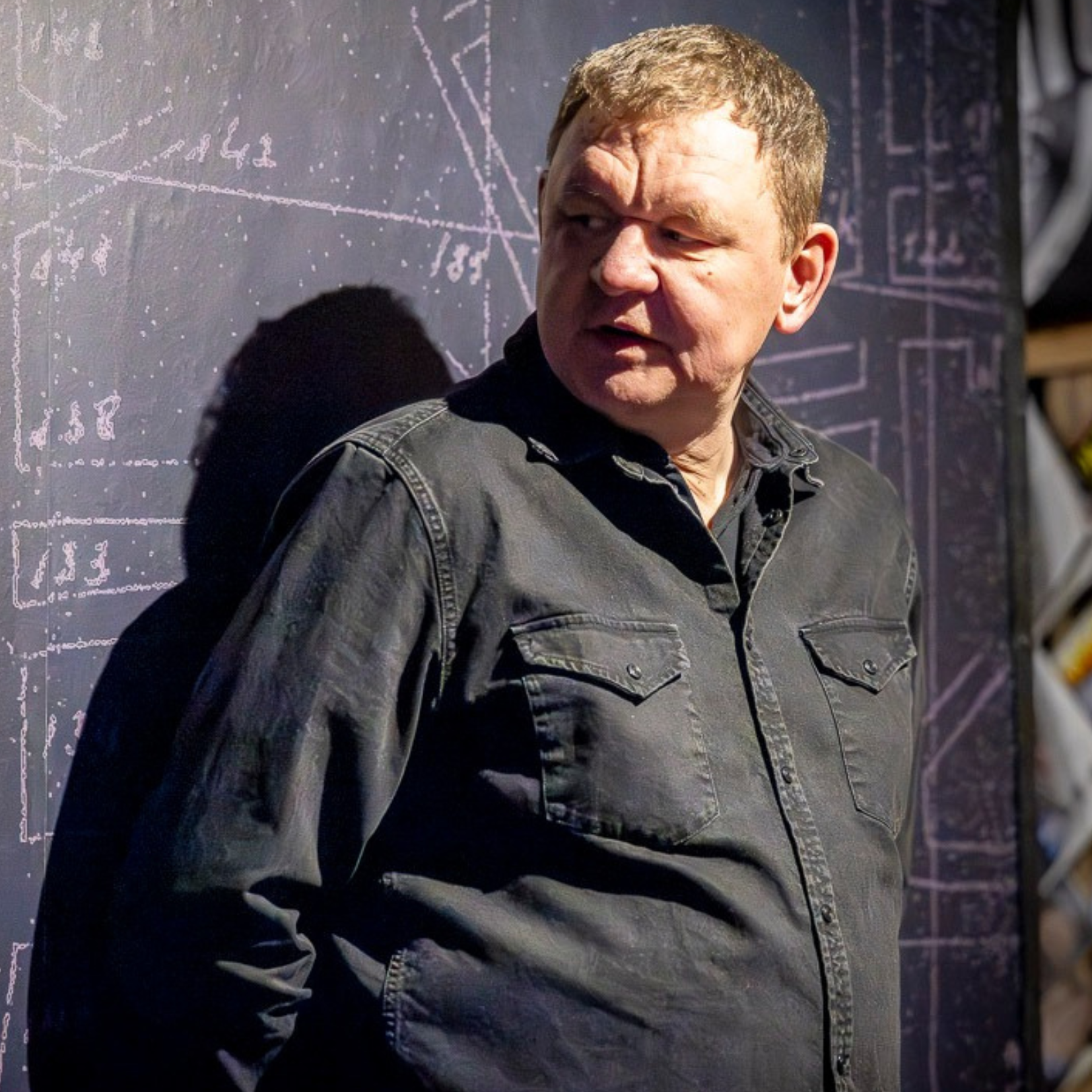 Krzysztof „Czarny” Krzyżanowski ubrana w ciemną koszulę z długim rękawem stoi bokiem przy tablicy z narysowanymi schematami i liniami, oświetlona punktowym światłem, na tle ciemnej ściany z technicznymi rysunkami.