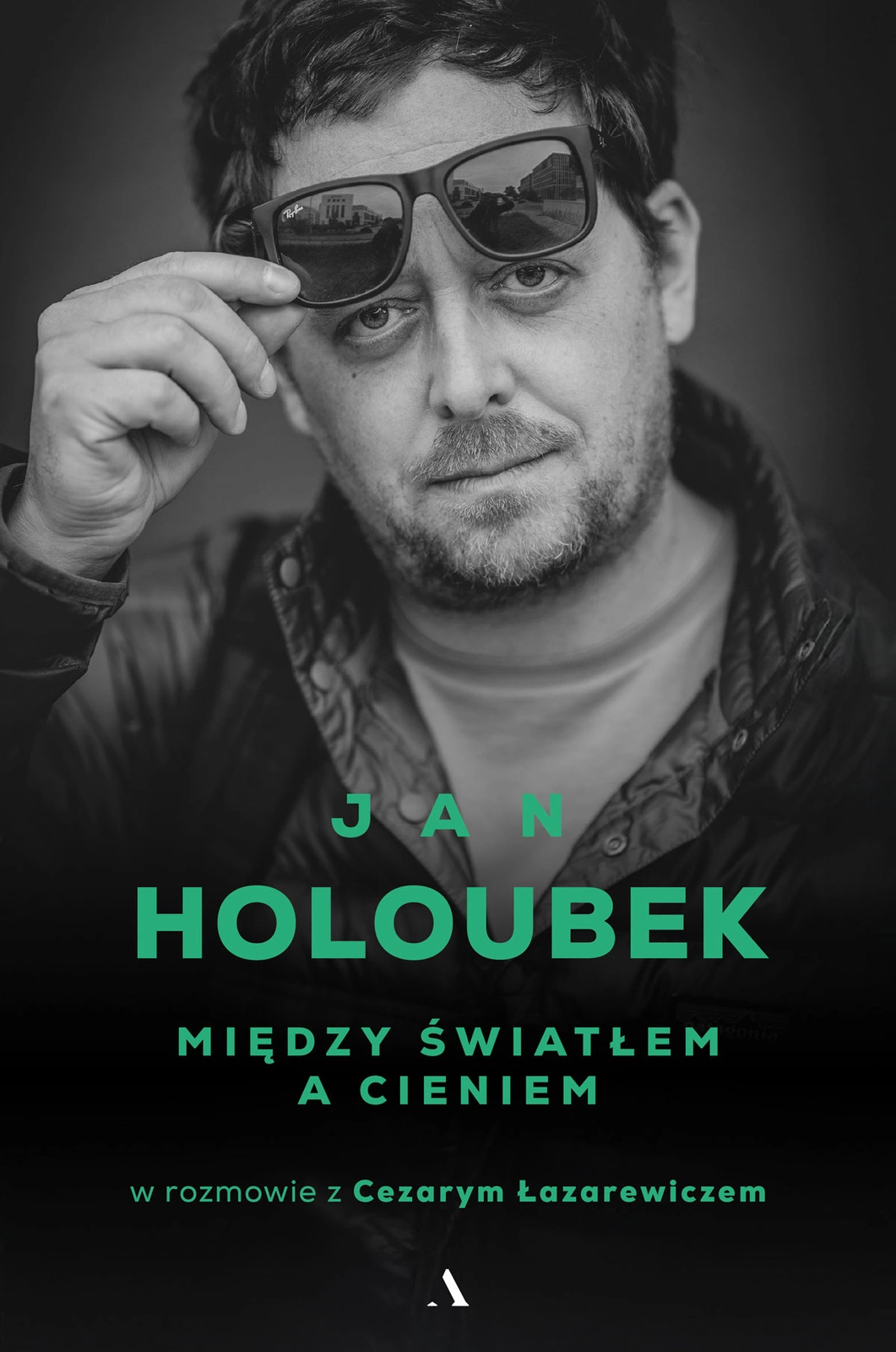 Okładka książki „Między światłem a cieniem” Jana Holoubka, w rozmowie z Cezarym Łazarewiczem. Czarno-białe zdjęcie portretowe mężczyzny w okularach przeciwsłonecznych, które unosi dłonią. Na dole czarny obszar z zielonymi napisami: „JAN HOLOUBEK”, „MIĘDZY ŚWIATŁEM A CIENIEM” oraz „w rozmowie z Cezarym Łazarewiczem”.