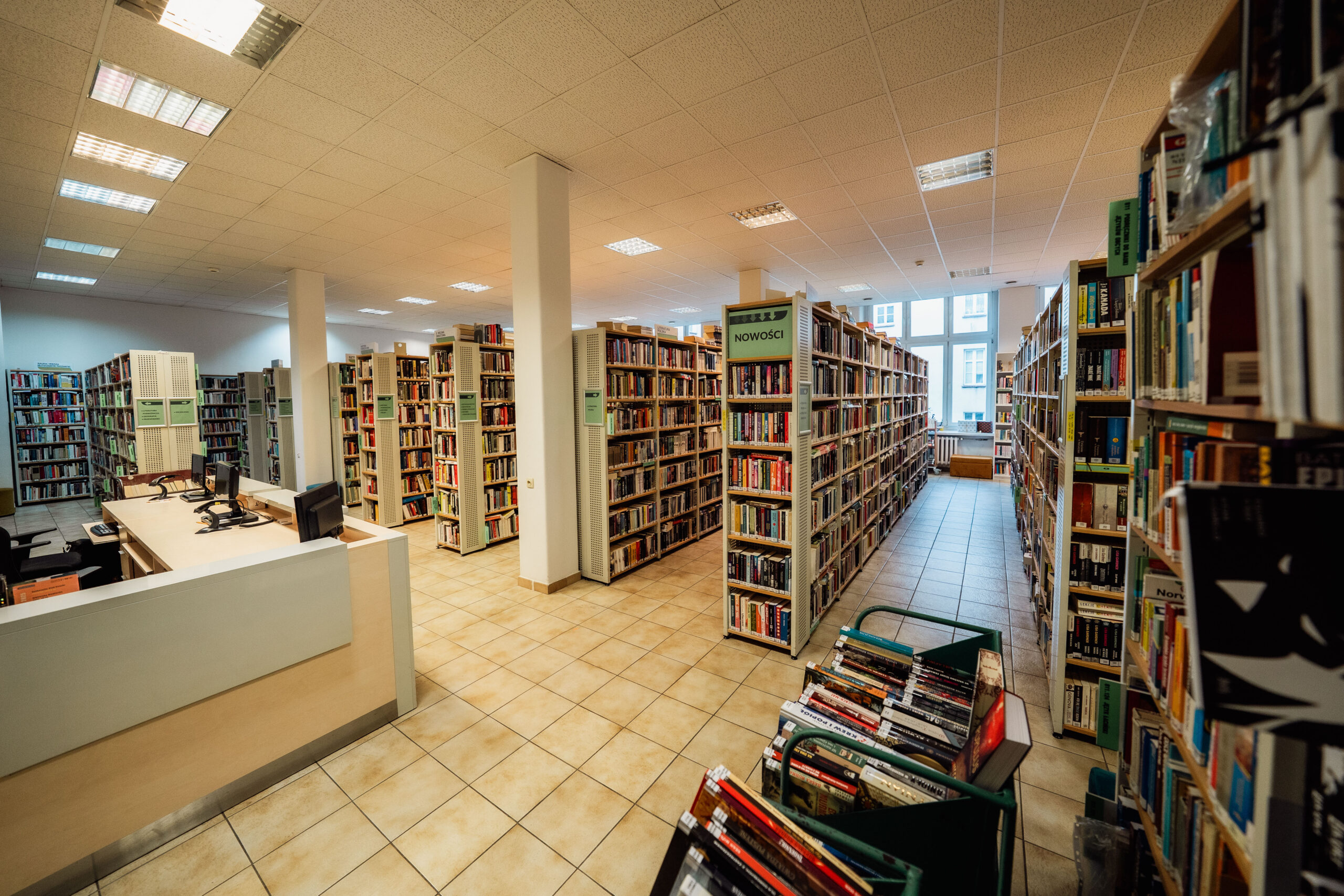 wypożyczalnia, otwarta przestrzeń, rzędy regałów z książkami, biblioteczna lada i wózek