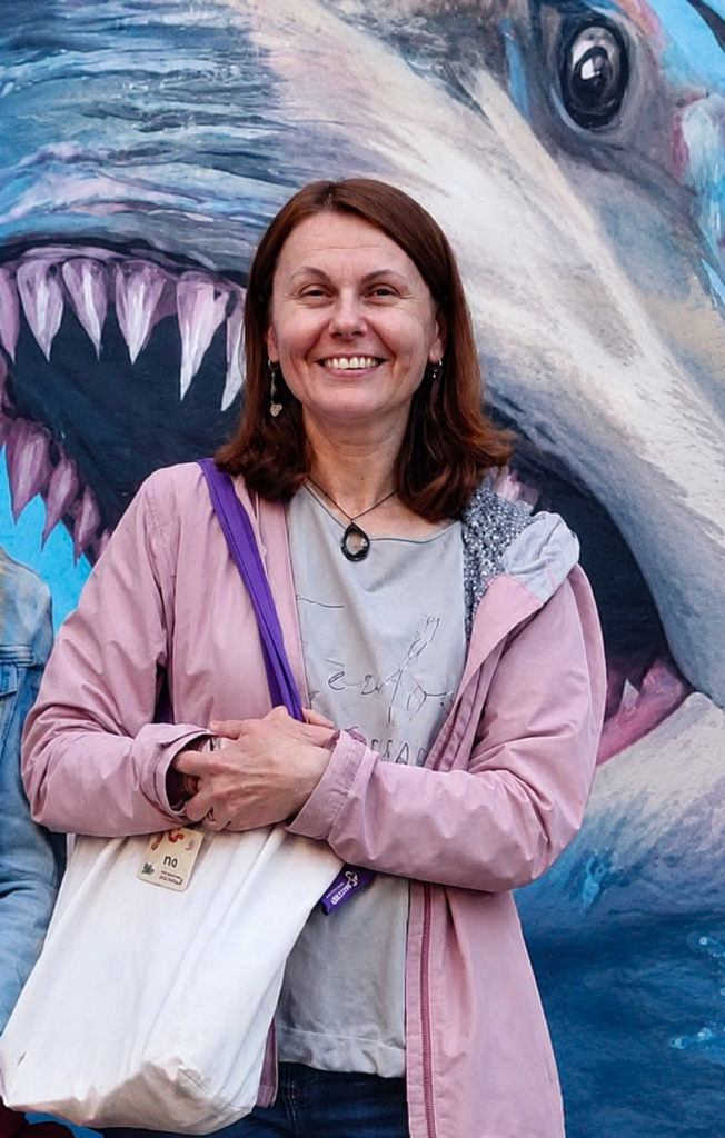 Elżbieta Mirowska w różowej kurtce i z białą torbą stoi na tle muralu przedstawiającego rekina wynurzającego się z wody