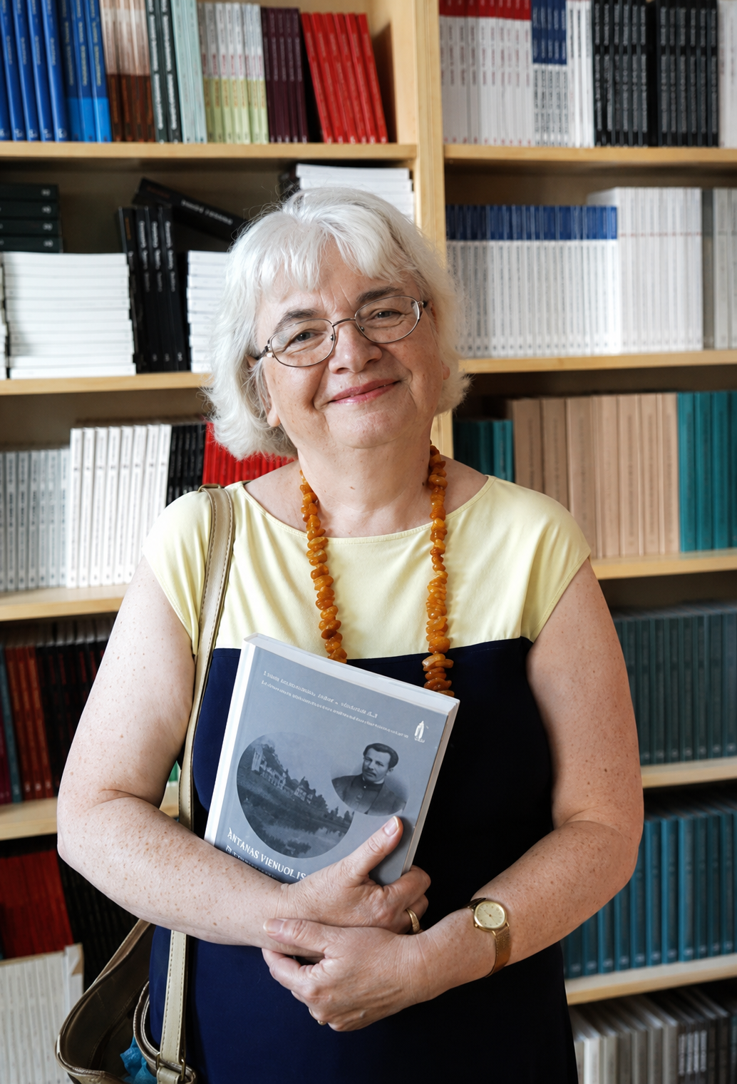 Małgorzata Łoboz pozuje z książką na tle regału z książkami. 
