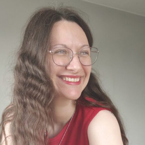 Zbliżeniowy portret kobiety z długimi, falowanymi włosami i okrągłymi okularami w cienkich oprawkach. Ubrana jest w czerwony bezrękawnik i ma na szyi delikatny naszyjnik. Zdjęcie wykonano we wnętrzu na jasnym, neutralnym tle. Głowa jest lekko zwrócona w bok, a na twarzy widoczny jest szeroki uśmiech.