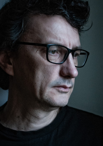 Zbliżenie twarzy mężczyzny w czarnych okularach. Patrzy w bok, poza kadr. Ma ciemne, lekko falowane włosy i poważny wyraz twarzy.