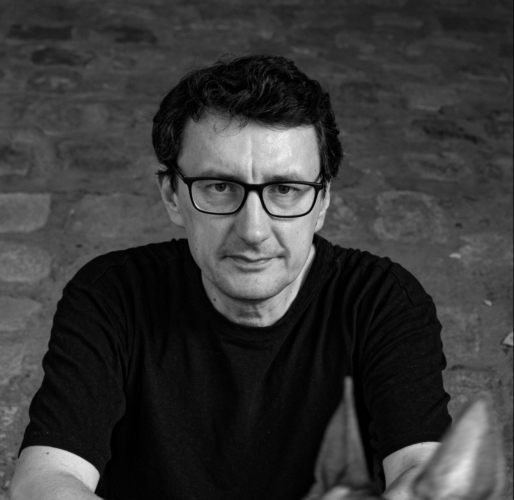 Czarno-białe zdjęcie mężczyzny w okularach i ciemnej koszulce. Siedzi z rękami opartymi przed sobą, patrząc prosto w obiektyw. W tle widoczny jest kamienny mur.