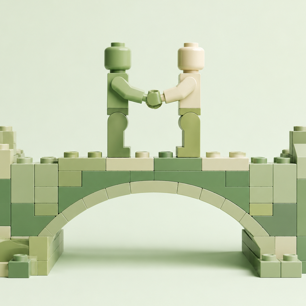 Minimalistyczna ilustracja 3D wykonana w stylistyce klocków LEGO. Na środku znajduje się niewielki most zbudowany z jasnych, zielonych i kremowych klocków. Na moście stoją naprzeciw siebie dwie uproszczone postacie i podają sobie dłonie. Całość utrzymana jest w spokojnej, pastelowej kolorystyce zieleni i beżu, na jasnym tle pozbawionym zbędnych elementów. Grafika symbolizuje dialog, spotkanie, współpracę oraz budowanie mostów między ludźmi.