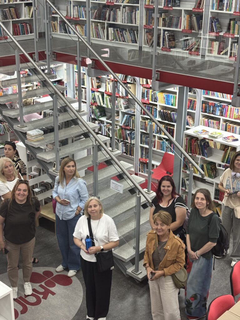 Widok wnętrza nowoczesnej biblioteki z antresolą i metalowymi schodami prowadzącymi na górny poziom. Między wysokimi regałami wypełnionymi książkami stoi grupa uczestników zwiedzania lub spotkania, którzy spoglądają w kierunku fotografa. Przestrzeń jest jasna i przestronna, z białymi regałami, szklanymi balustradami oraz czerwonymi akcentami kolorystycznymi. Na półkach widoczne są oznaczenia działów tematycznych. Zdjęcie wykonano z wyższej kondygnacji, dzięki czemu widać układ biblioteki oraz centralnie usytuowane schody.