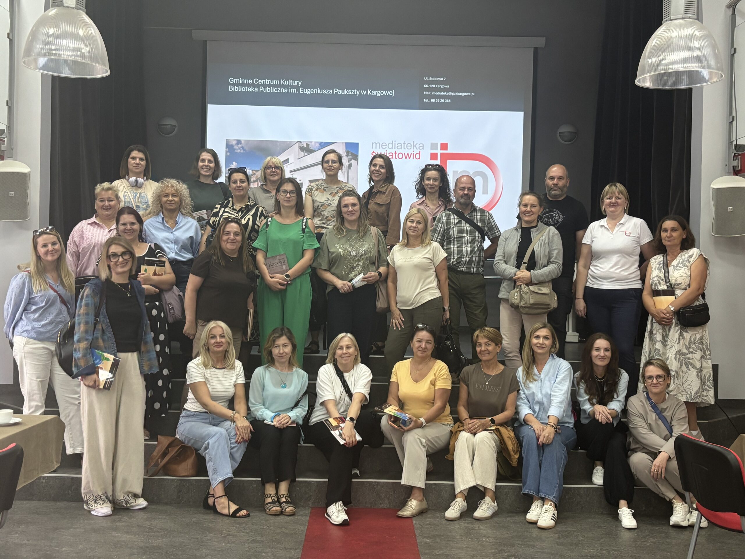 Grupowe zdjęcie około 30 uczestników wydarzenia stojących i siedzących na schodach w sali konferencyjnej. W tle widoczny jest ekran projekcyjny z napisem „Gminne Centrum Kultury Biblioteka Publiczna im. Eugeniusza Paukszty w Kargowej” oraz logo „Mediateka Światowid”. Uczestnicy pozują do wspólnego zdjęcia, część trzyma materiały lub książki. Wnętrze oświetlają duże wiszące lampy, a przez środek schodów biegnie czerwony pasek.