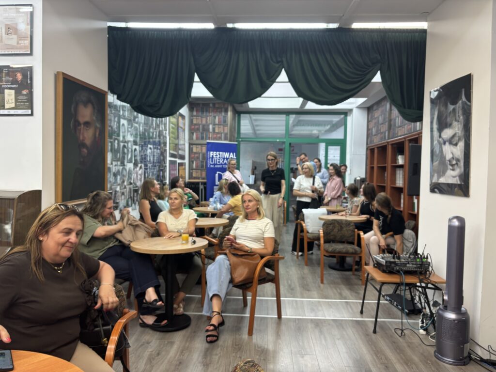 Wąskie wnętrze biblioteki lub czytelni z kilkunastoma osobami siedzącymi przy okrągłych stolikach i rozmawiającymi przed rozpoczęciem wydarzenia. Na ścianach znajdują się portrety, fotografie i regały z książkami. W głębi sali widoczny jest baner z napisem „Festiwal Literacki”, a po prawej stronie sprzęt nagłośnieniowy. Pomieszczenie jest jasno oświetlone, z ciemnozielonymi kotarami zawieszonymi pod sufitem.